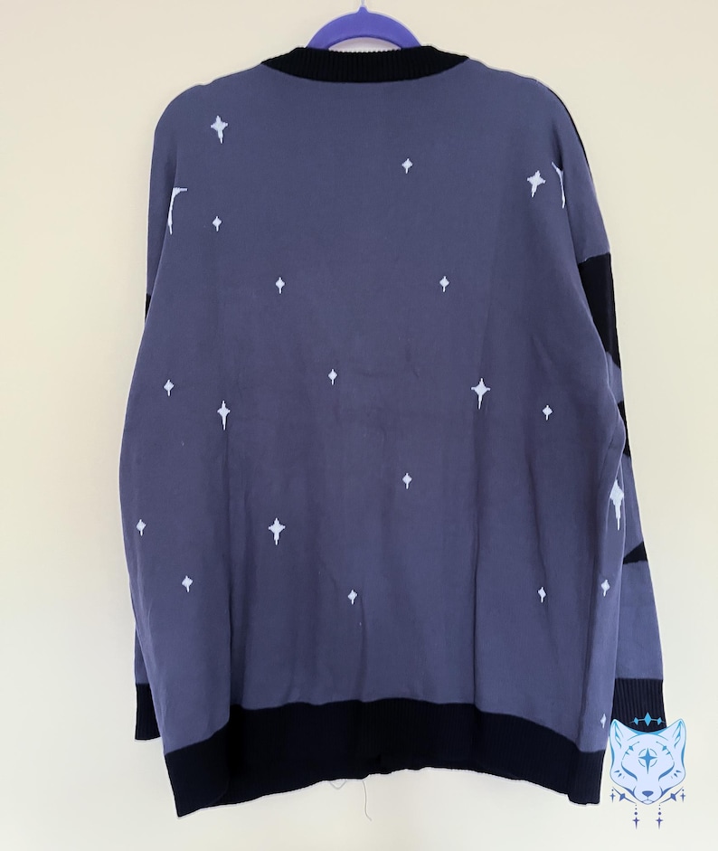 May include: A dark blue sweater with white star patterns. The sweater has a black collar, cuffs, and hem. The back of the sweater is visible, hanging on a purple hanger. The sweater is a knitwear item.