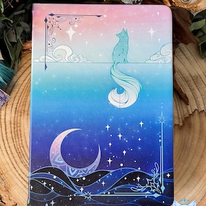 May include: A blue and pink notebook with a white fox with a long tail and a crescent moon design. The notebook has a gradient background with stars and clouds.