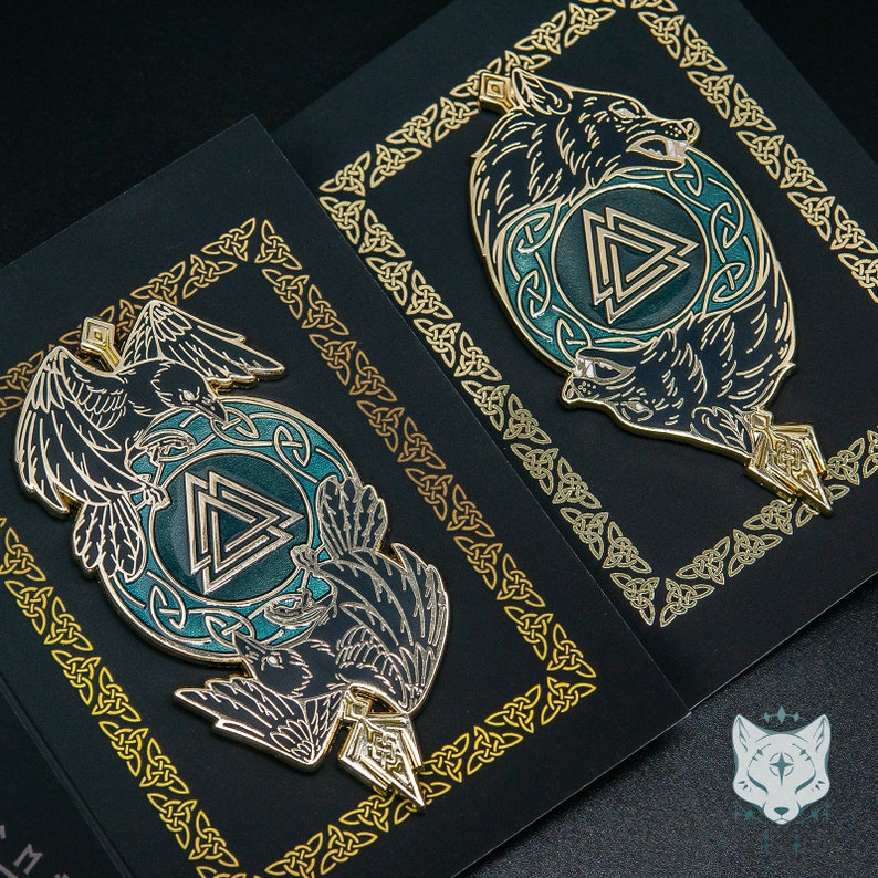 May include: Two enamel pins with gold accents on a black background. One pin features a raven and a wolf, the other features a wolf and a raven. Both pins have a green and blue design with a triangular symbol.