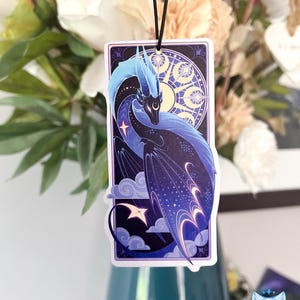 Pu&ograve; includere: Un drago blu e viola con stelle e lune sulle ali, su uno sfondo blu scuro con nuvole. Il drago &egrave; in una cornice bianca con un bordo viola.