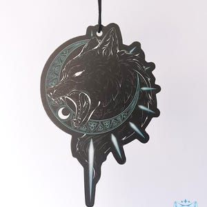 May include: Black and white car air freshener featuring a wolf howling at the moon. The wolf is encircled by a circle of Celtic knots and the moon is crescent shaped. The air freshener is suspended from a black string.