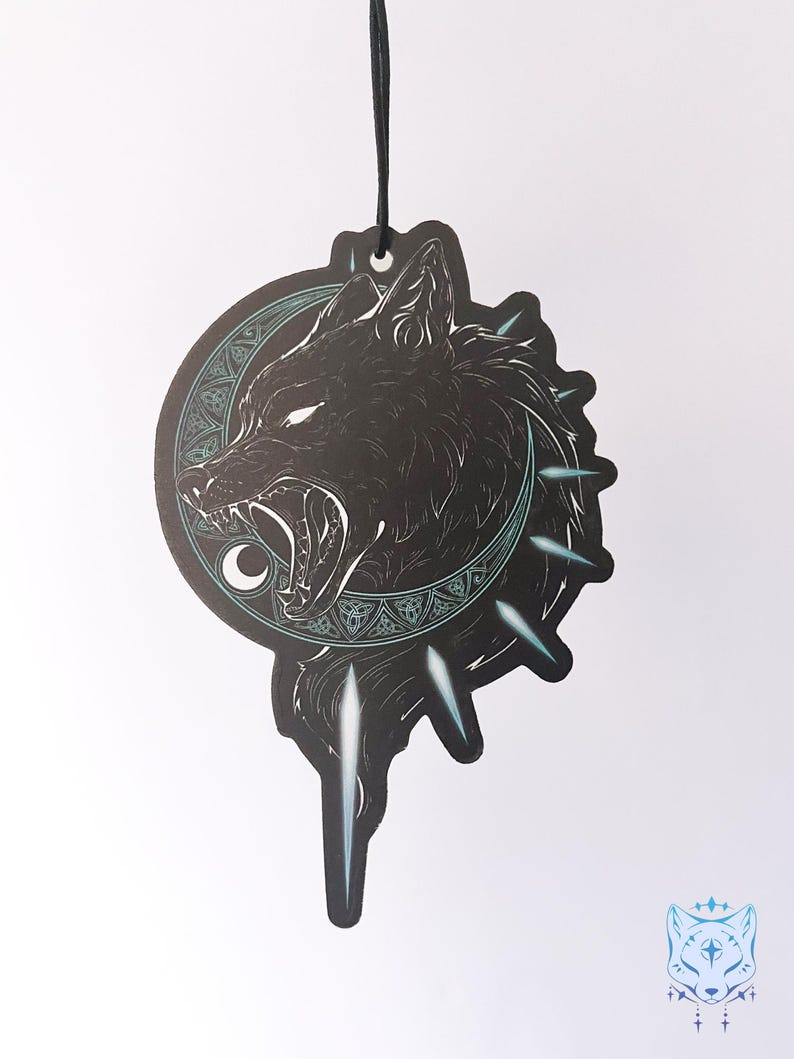 May include: Black and white car air freshener with a wolf howling at the moon. The wolf is surrounded by a circle of Celtic knots and the moon is crescent shaped. The air freshener is hanging from a black string.