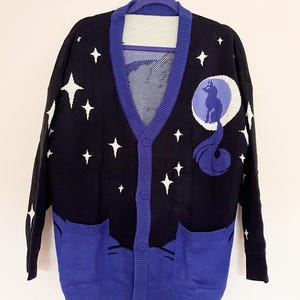 May include: Black cardigan sweater with a celestial design. The sweater features a navy blue background with white stars and a moon and wolf graphic. The trim and pockets are a vibrant blue. The sweater has a button-up front and two front pockets.