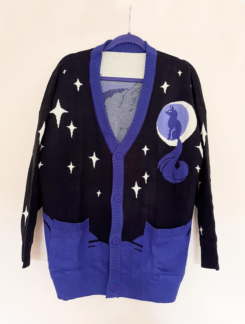 May include: Black cardigan sweater with a celestial design. The sweater features a navy blue background with white stars and a moon and wolf graphic. The trim and pockets are a vibrant blue. The sweater has a button-up front and two front pockets.
