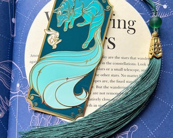 May include: A teal blue and gold bookmark featuring a stylized fox with a white rabbit. The bookmark has a tassel and a gold charm.