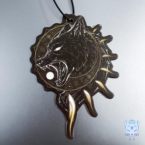 May include: Black and gold car air freshener with a wolf's head design. The wolf is snarling and has a white circle in its mouth. The design is surrounded by a gold circle with intricate patterns.