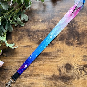 Op de afbeelding: Een blauwe en roze lanyard met een galaxy print en witte sterren. De lanyard heeft een zilveren metalen clip.