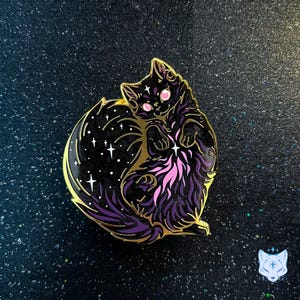 Puede incluir: Un pin de esmalte dorado con un gato negro enroscado en una bola con un diseño de cielo nocturno estrellado. El gato tiene ojos rosas y una cola morada y negra.