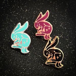 May include: Three enamel pins featuring a stylized rabbit design. The rabbits are curled up in a sleeping position. The pins are in three different colors: teal, burgundy, and black.