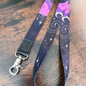 Puede incluir: Un cord&oacute;n de tela con un patr&oacute;n celestial en morado oscuro y negro que incluye estrellas blancas, nubes arremolinadas y lunas crecientes. La correa est&aacute; unida a una leng&uuml;eta de cuero negro con un emblema de cabeza de lobo y un mosquet&oacute;n giratorio de metal plateado.