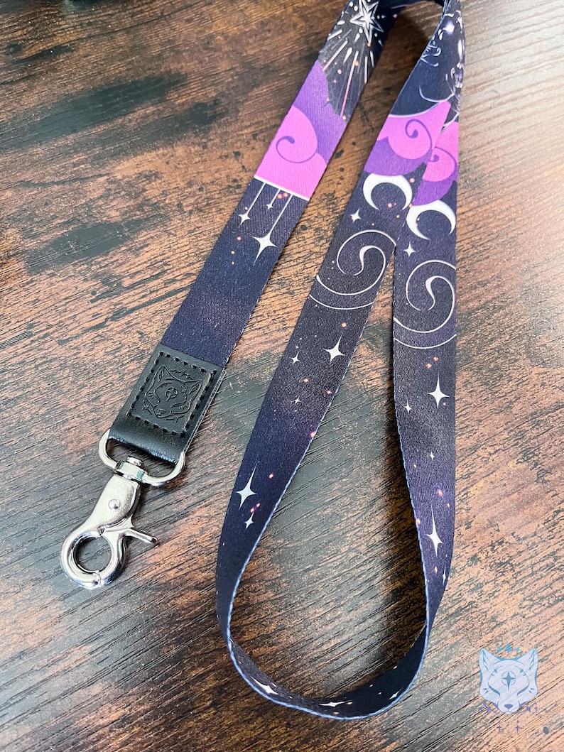 Puede incluir: Un cord&oacute;n de tela con un patr&oacute;n celestial en morado oscuro y negro que incluye estrellas blancas, nubes arremolinadas y lunas crecientes. La correa est&aacute; unida a una leng&uuml;eta de cuero negro con un emblema de cabeza de lobo y un mosquet&oacute;n giratorio de metal plateado.