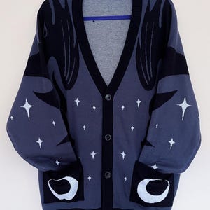 May include: A button-down cardigan in a dark blue hue, featuring black accents and white star and crescent moon designs. The sweater has a V-neck, long sleeves, and two front pockets. The cardigan is hanging on a blue hanger.