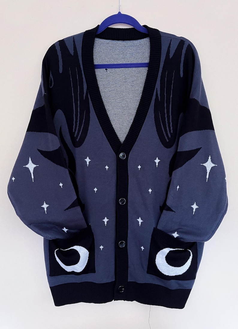 May include: A button-down cardigan in a dark blue hue, featuring black accents and white star and crescent moon designs. The sweater has a V-neck, long sleeves, and two front pockets. The cardigan is hanging on a blue hanger.