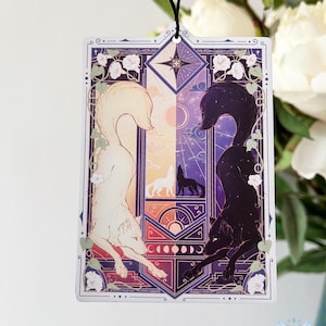 May include: Rectangular air freshener featuring a symmetrical design of a white and black wolf. The artwork includes a celestial theme with a star, moon phases, and floral accents. A black string is attached for hanging.