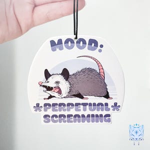 May include: White car air freshener with a cartoon illustration of a possum with the text "MOOD: *PERPETUAL* SCREAMING"