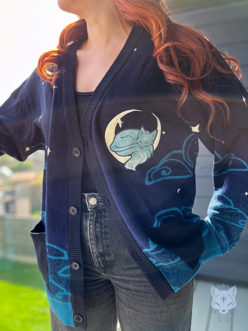 May include: Navy blue cardigan with a celestial design. Features a sleeping fox on a crescent moon, clouds, and stars. The cardigan has a button-up front, pockets, and a light blue trim. The sleeves and pockets have a gradient blue design.