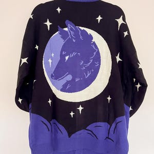 May include: Black cardigan sweater with a celestial design. The back features a purple wolf head inside a white crescent moon, surrounded by white stars on a black background. The bottom and collar are purple.
