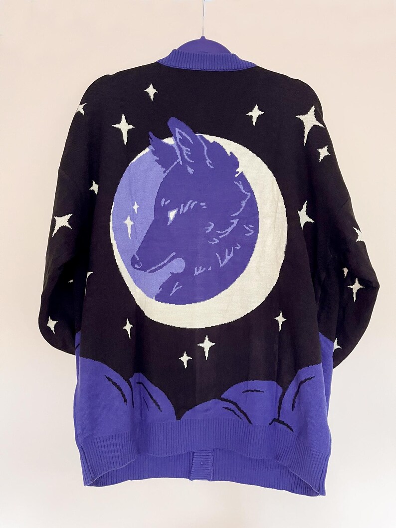 May include: Black cardigan sweater with a celestial design. The back features a purple wolf head inside a white crescent moon, surrounded by white stars on a black background. The bottom and collar are purple.