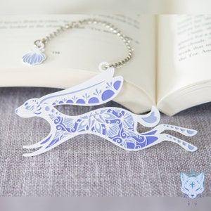 May include: A white metal bookmark shaped like a running hare with a purple floral design. The bookmark has a silver chain attached to a small shell charm.