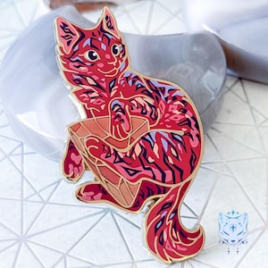 Puede incluir: Un pin de esmalte rojo y rosa de un gato enroscado en una forma geométrica rosa y naranja. El gato tiene un patrón rayado y una cara blanca con ojos negros.