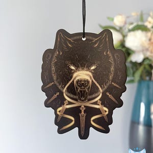 以下が含まれることがあります： 光る目と牙を持つ狼の頭が描かれた、黒と金の車の芳香剤。狼は、短剣の円と金色のリボンで囲まれています。
