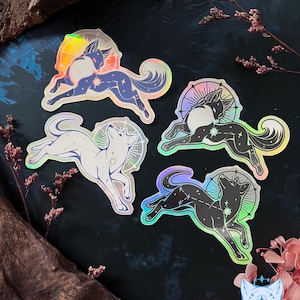 Puede incluir: Cuatro pegatinas holográficas con un diseño de lobo estilizado y un patrón de constelación. Los lobos son de color blanco, negro y azul, con un efecto de halo arcoíris. Las pegatinas están sobre un fondo oscuro con detalles florales.
