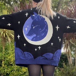 May include: Black sweater with a blue wolf and moon design. The sweater features a large wolf silhouette inside a crescent moon, surrounded by white stars. The bottom hem and cuffs are a deep blue color.