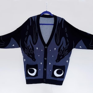 May include: A black and blue cardigan sweater with a dragon design. The sweater has a V-neck, button closure, and two pockets with a crescent moon design. White stars are scattered across the blue background.