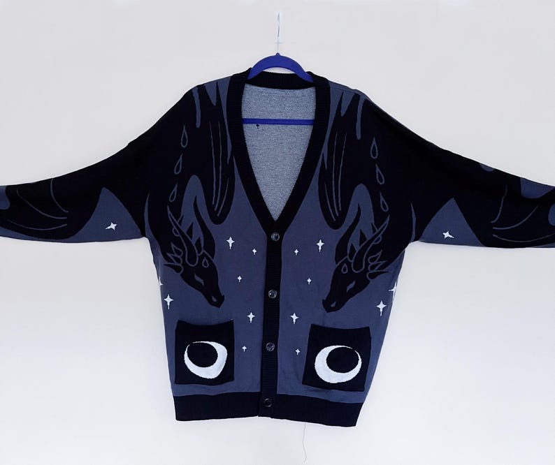 May include: A black and blue cardigan sweater with a dragon design. The sweater has a V-neck, button closure, and two pockets with a crescent moon design. White stars are scattered across the blue background.