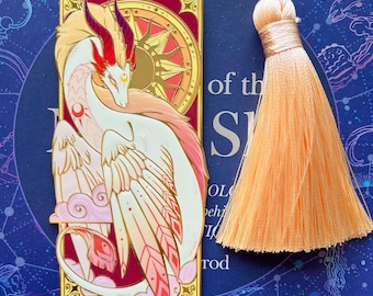 Puede incluir: Marcapáginas de metal dorado con un diseño de dragón blanco y rosa con cuernos rojos y una borla. El marcapáginas tiene forma rectangular con un diseño de rosa de los vientos y una borla color melocotón.