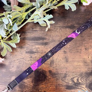 Puede incluir: Un cord&oacute;n de tela con un patr&oacute;n celestial en morado oscuro y negro, que presenta ilustraciones en blanco de estrellas, una luna creciente y la silueta de un gato. La cinta incluye un mosquet&oacute;n giratorio de metal plateado con una leng&uuml;eta de piel sint&eacute;tica.