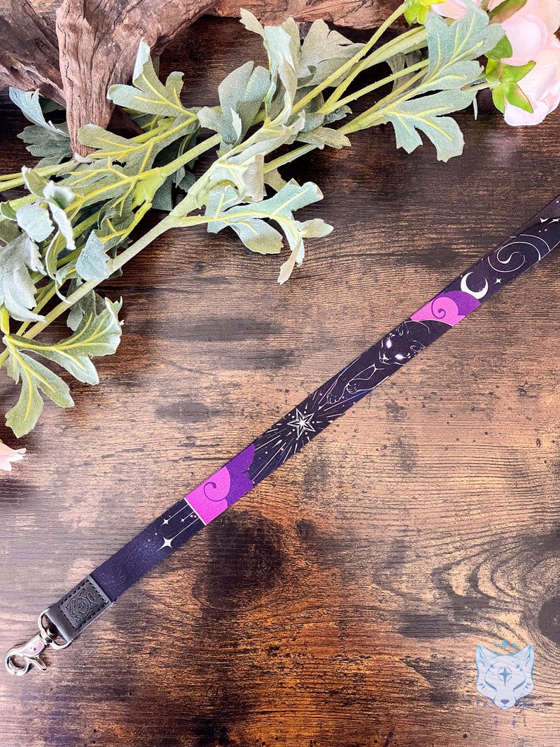 Puede incluir: Un cord&oacute;n de tela con un patr&oacute;n celestial en morado oscuro y negro, que presenta ilustraciones en blanco de estrellas, una luna creciente y la silueta de un gato. La cinta incluye un mosquet&oacute;n giratorio de metal plateado con una leng&uuml;eta de piel sint&eacute;tica.