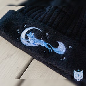 May include: Black knit beanie with an embroidered design of a blue fox jumping over a crescent moon with stars.