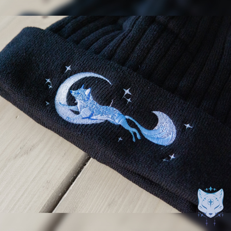 May include: Black knit beanie with an embroidered design of a blue fox jumping over a crescent moon with stars.
