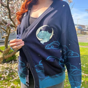 May include: Navy blue cardigan with a cat and moon design. The cardigan features a light blue trim, pockets, and a button-up front. The design includes clouds and stars. The cardigan is made of a soft material.