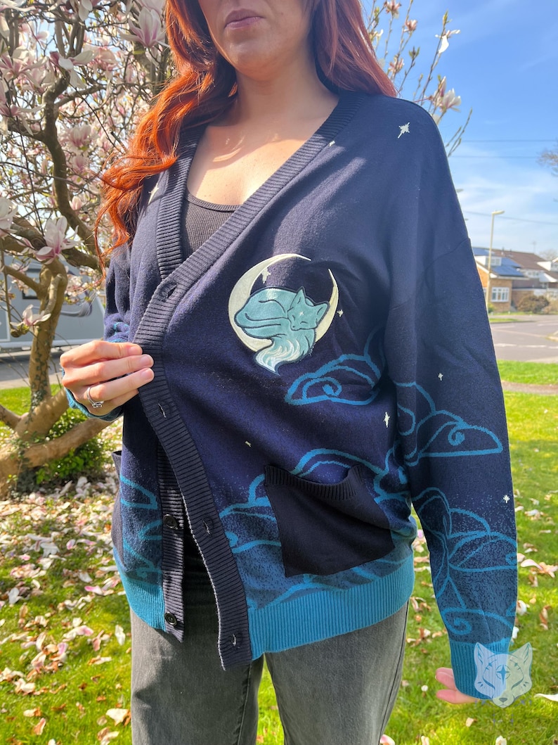 May include: Navy blue cardigan with a cat and moon design. The cardigan features a light blue trim, pockets, and a button-up front. The design includes clouds and stars. The cardigan is made of a soft material.