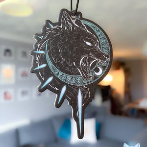 May include: Black and white car air freshener with a wolf howling at a crescent moon. The wolf is surrounded by a circle of blue and white geometric designs. The air freshener is hanging from a black string.