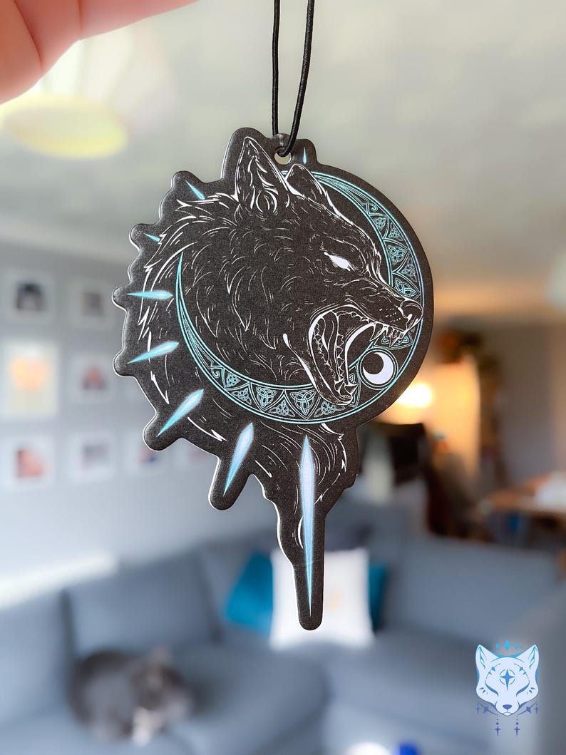 May include: Black and white car air freshener with a wolf howling at a crescent moon. The wolf is surrounded by a circle of blue and white geometric designs. The air freshener is hanging from a black string.
