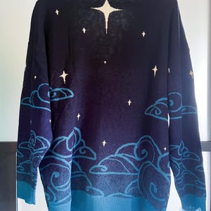 May include: Navy blue sweater with a celestial design. The sweater features a pattern of white stars and stylized blue clouds against a dark blue background. The cuffs and hem are a lighter shade of blue.