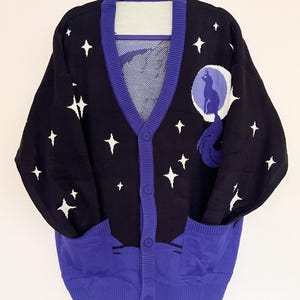 May include: Black cardigan sweater with a celestial design. The sweater features white star patterns, a purple trim, and a large graphic of a cat on the moon. It has two front pockets and is buttoned up.