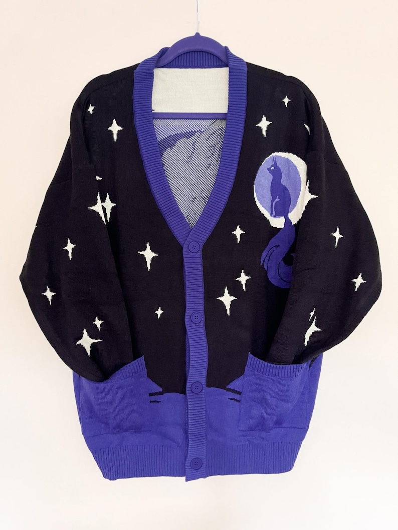 May include: Black cardigan sweater with a celestial design. The sweater features white star patterns, a purple trim, and a large graphic of a cat on the moon. It has two front pockets and is buttoned up.