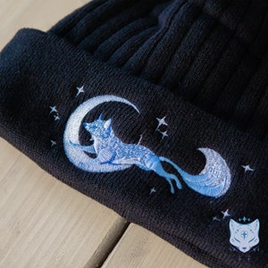 May include: Black knit beanie with a blue embroidered design of a fox curled around a crescent moon with stars.