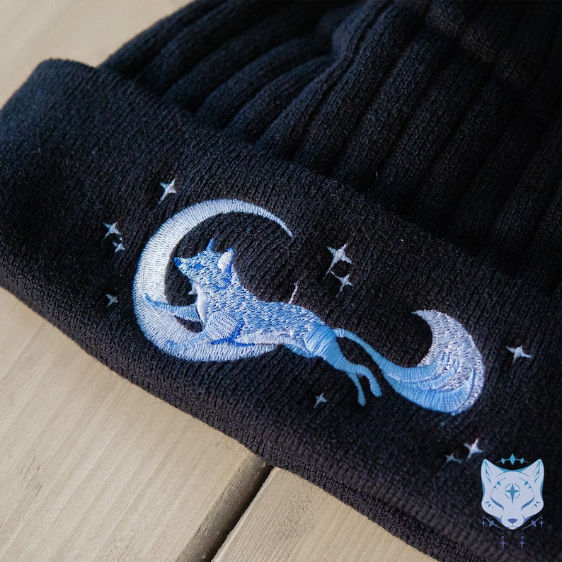 May include: Black knit beanie with a blue embroidered design of a fox curled around a crescent moon with stars.