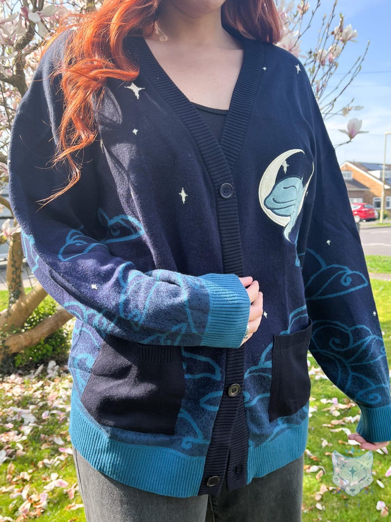 May include: Navy blue cardigan with a celestial design. The sweater features a V-neck, button closure, and two front pockets. The design includes a moon and a creature, clouds, and stars in shades of blue and white.