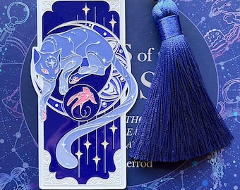 Puede incluir: Un marcapáginas de metal blanco con un diseño azul y blanco de un gato enroscado alrededor de una luna creciente. El gato tiene un pez rosa en la boca. El marcapáginas tiene un borla azul.
