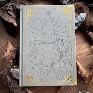 Könnte beinhalten: Ein graues Hardcover-Buch mit einer geprägten Illustration eines heulenden Wolfes, umgeben von Rosen und einer Kette. Das Cover-Design ist mit Goldfolienakzenten und dekorativen Ecken verziert, mit einem Sternsymbol über dem Wolf.