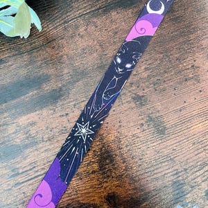 Puede incluir: Un cord&oacute;n de tela con fondo oscuro y un patr&oacute;n repetitivo de un gato negro con ojos brillantes, estrellas blancas, lunas crecientes y formas de nubes moradas. El dise&ntilde;o incluye elementos celestiales y motivos c&oacute;smicos estilizados.