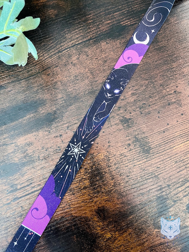Puede incluir: Un cord&oacute;n de tela con fondo oscuro y un patr&oacute;n repetitivo de un gato negro con ojos brillantes, estrellas blancas, lunas crecientes y formas de nubes moradas. El dise&ntilde;o incluye elementos celestiales y motivos c&oacute;smicos estilizados.