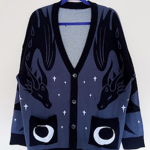 May include: A dark blue cardigan with a V-neck and button closure. The sweater features a dragon design on the front and sleeves, with white stars and crescent moon pockets. The trim and cuffs are black.