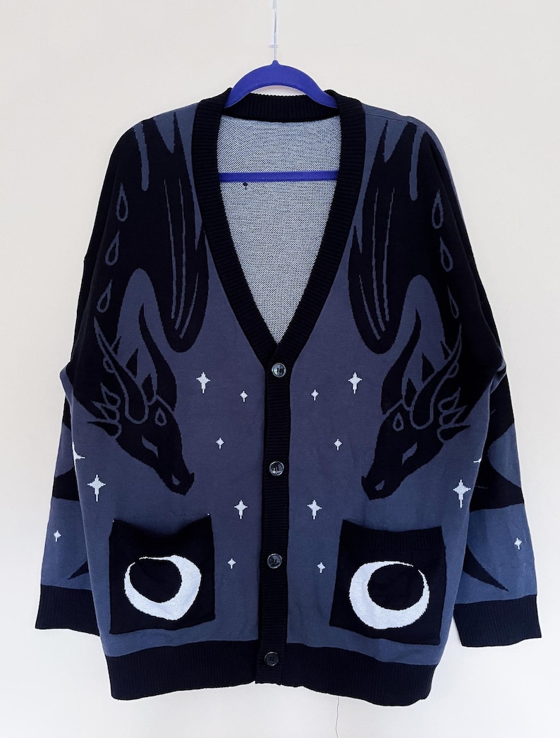May include: A dark blue cardigan with a V-neck and button closure. The sweater features a dragon design on the front and sleeves, with white stars and crescent moon pockets. The trim and cuffs are black.